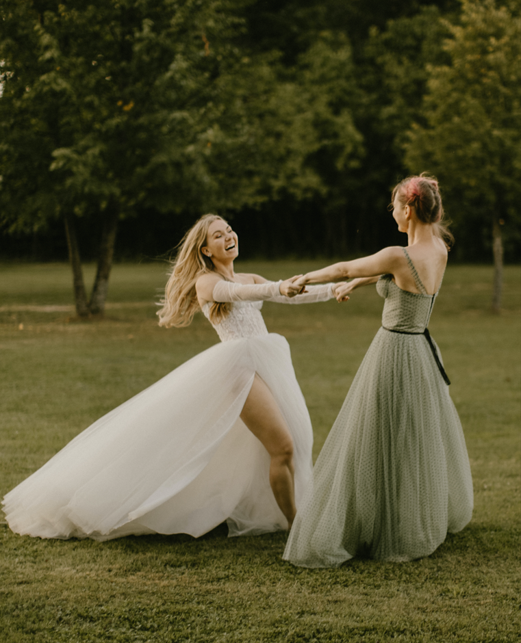 dvi moterys su balta ir žalia suknele sukasi susikibusios rankomis