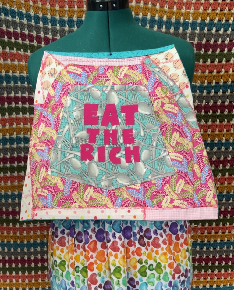 An Apron art piece that says EAT THE RICH in pink letters over utensils fabric accented with pig fab