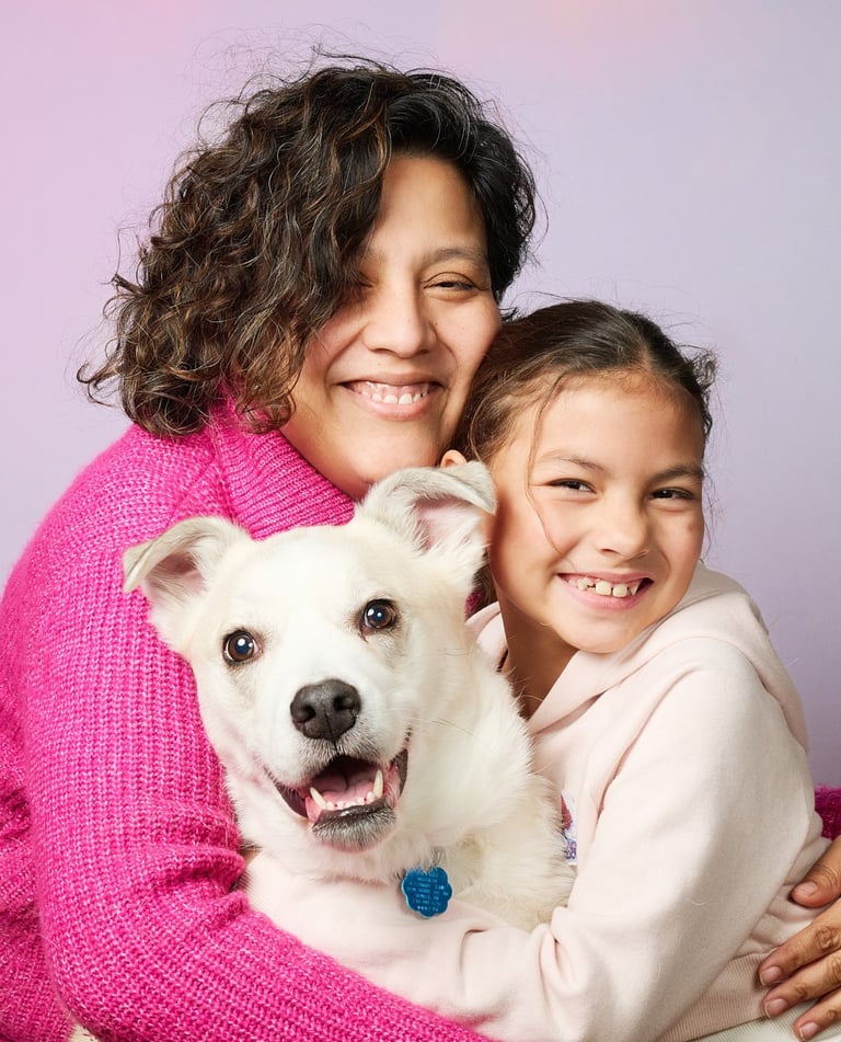 A smiling mother and daughter hugging their happy white pet dog against a purple background.