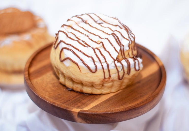 Rollo de canela recién horneado con glaseado blanco y un chorrito de chocolate sobre un pedestal