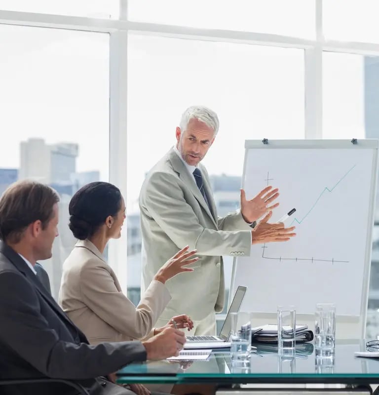 A man in a suit presents a rising graph on a flip chart to two colleagues in a bright conference room.