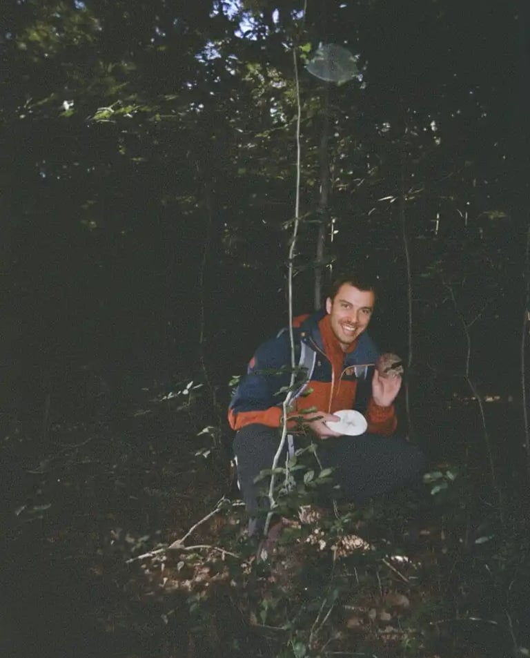 Smiling hiker in a red and blue jacket crouches in a dark forest holding a mushroom