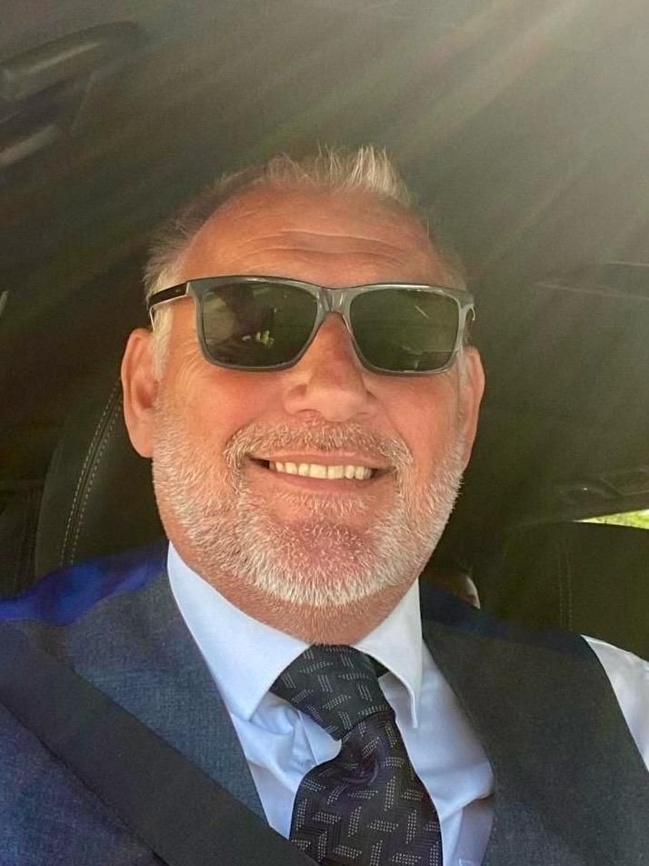 Nigel, smiling with a grey beard wearing sunglasses, a grey waistcoat, and a patterned tie.