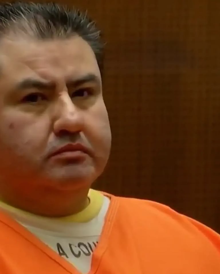 A man in an orange prison jumpsuit sits in a courtroom during a legal hearing.