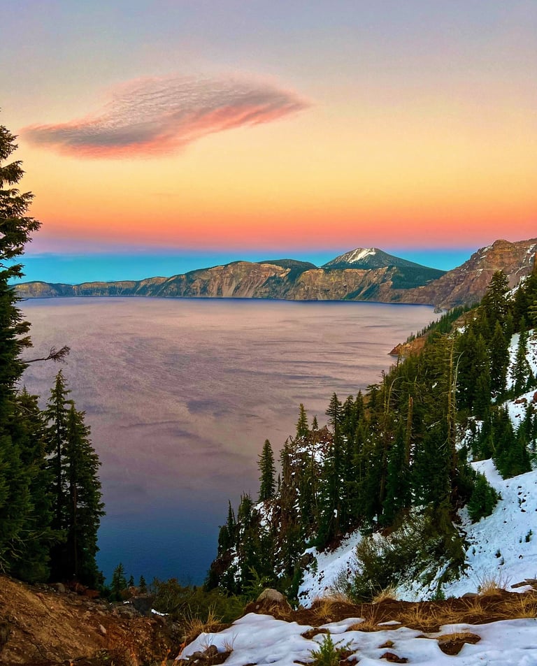 Sunset over Crater Lake National Park with snow-covered slopes and a pink sky over the deep blue caldera.