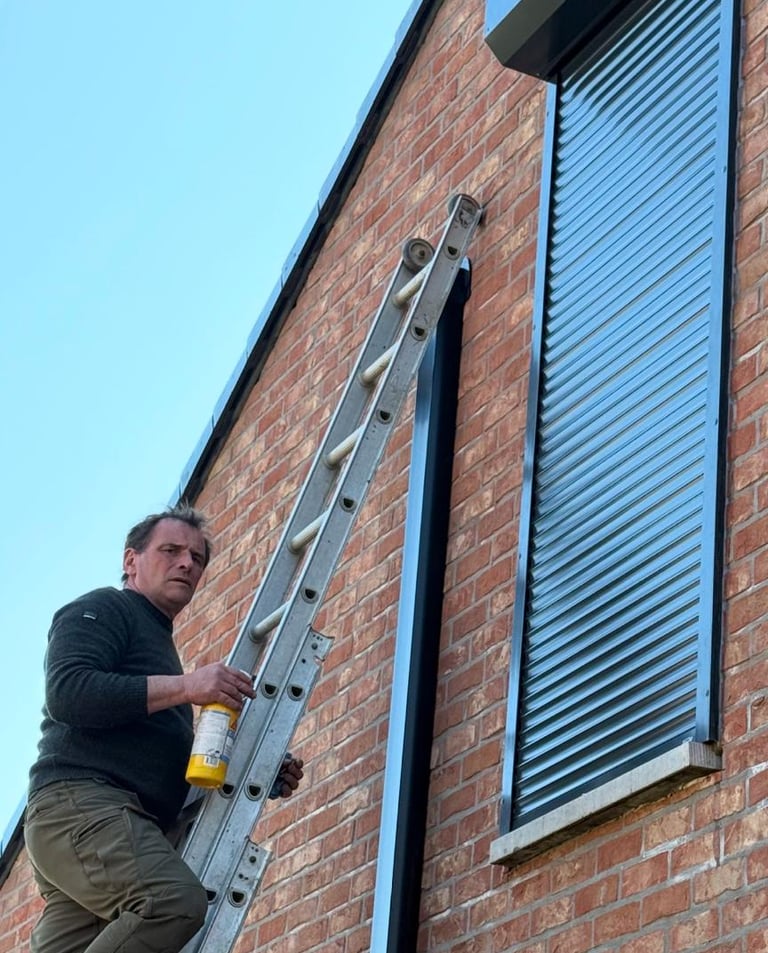 A technician on a ladder installs modern black security roller shutters on a brick residential house.