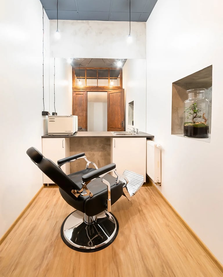 Mały gabinet z czarnym fotelem fryzjerskim, lustrem, umywalką i białymi ścianami.