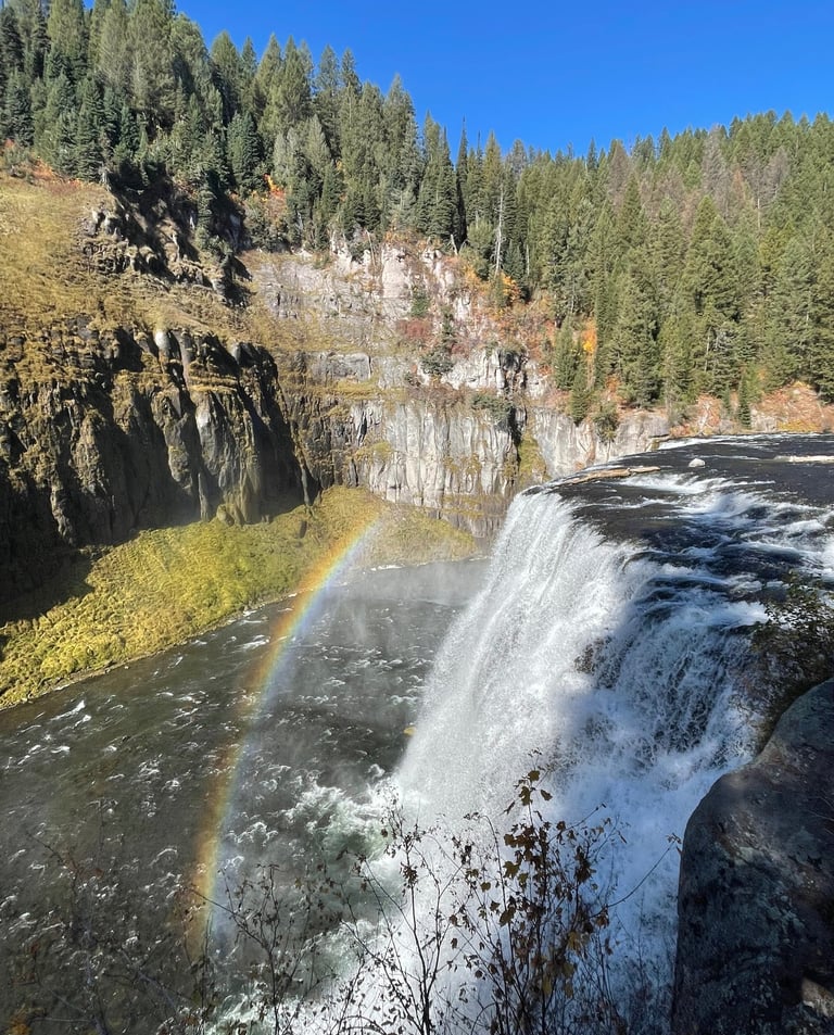 Mesa Falls cascading into a canyon with a vibrant rainbow over the water and evergreen forest.