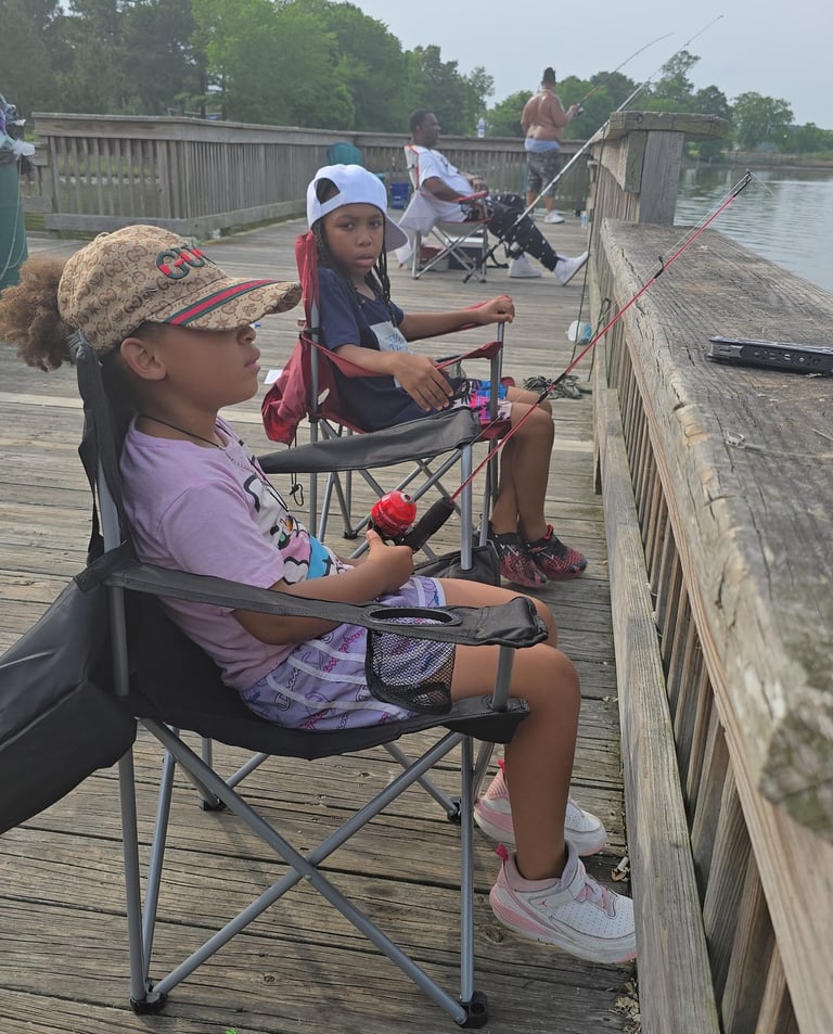 Two young girls sitting in folding chairs fishing from a wooden pier on a cloudy day.
