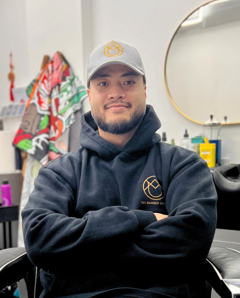 Yau, professional barber and founder of YAU Barber Studio, in his modern barber chair wearing branded hoodie and cap.