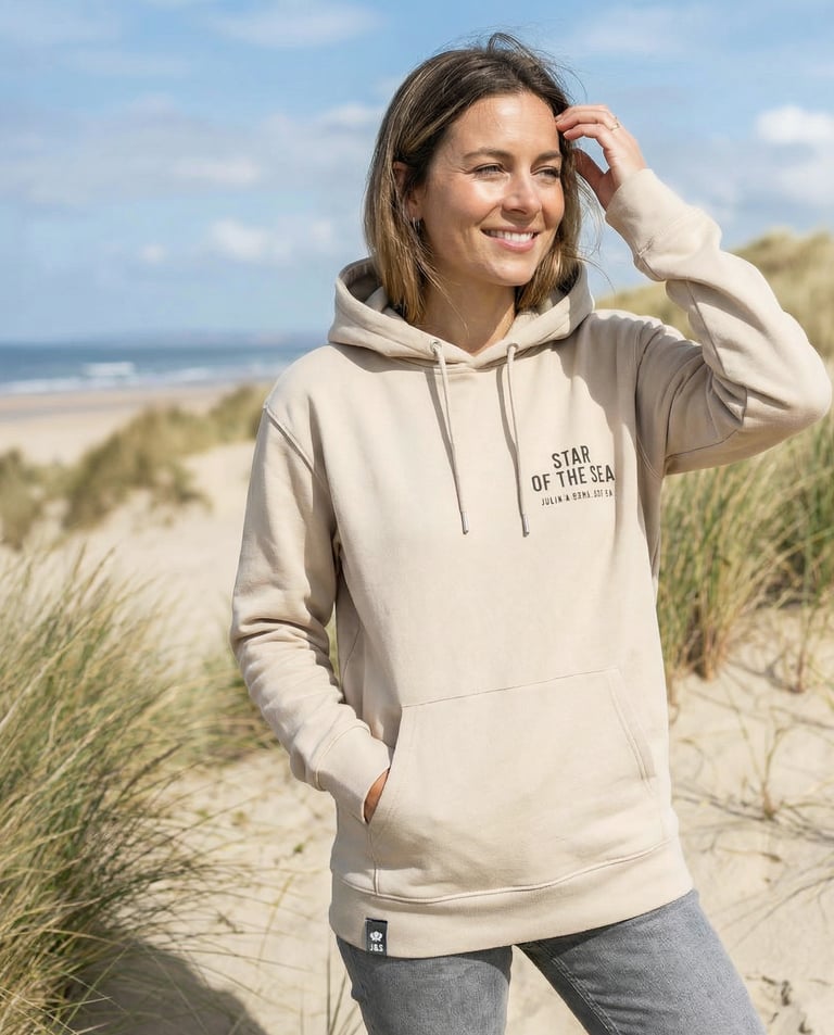 Smiling woman wearing a beige hoodie on a sandy beach with grassy dunes in the background.