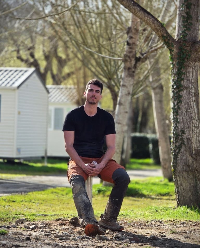 A man in workwear sitting at a sunny campsite with mobile homes and trees in the background.