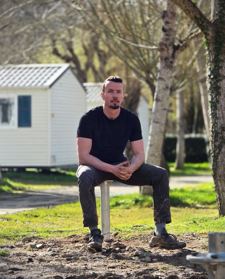 Man in black t-shirt sitting outdoors at a campground with mobile homes and trees in the background.