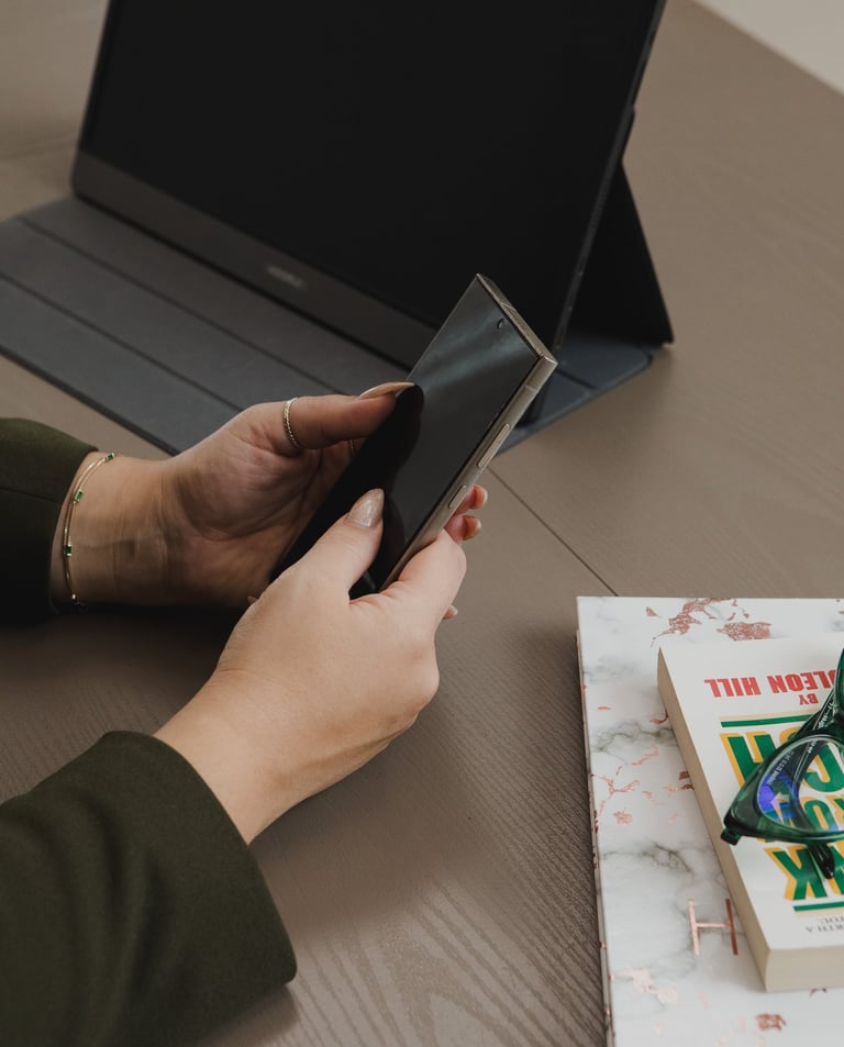 Adriana holding a smartphone while working at a desk with a laptop, tablet, and books.