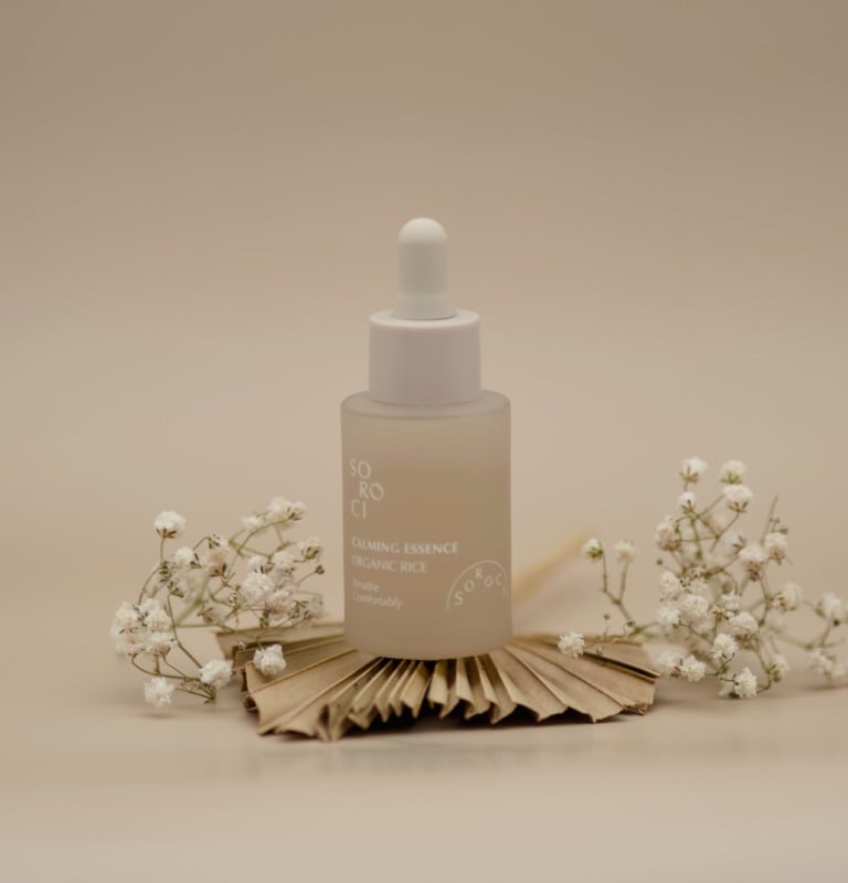 Sticluță de esență calmantă, cu pipetă albă, așezată central pe fundal bej, decorată cu flori albe delicate