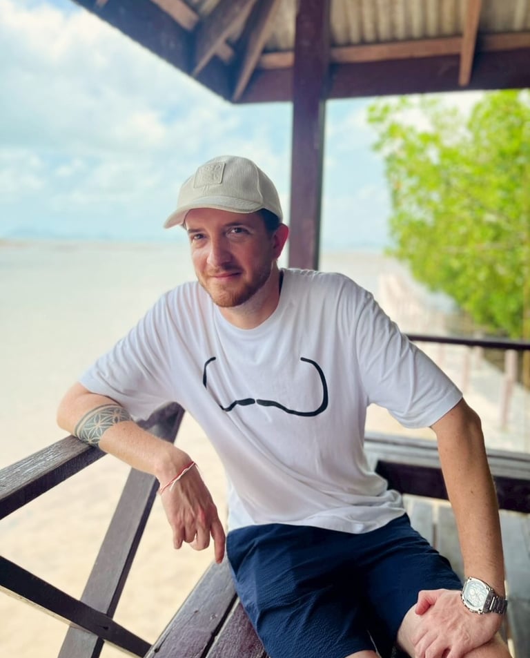 Unicris wearing a white t-shirt and baseball cap sitting on a wooden balcony at a tropical beach.
