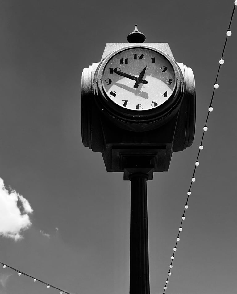 A black-and-white photograph of a vintage street clock mounted on a tall post against a clear sky.