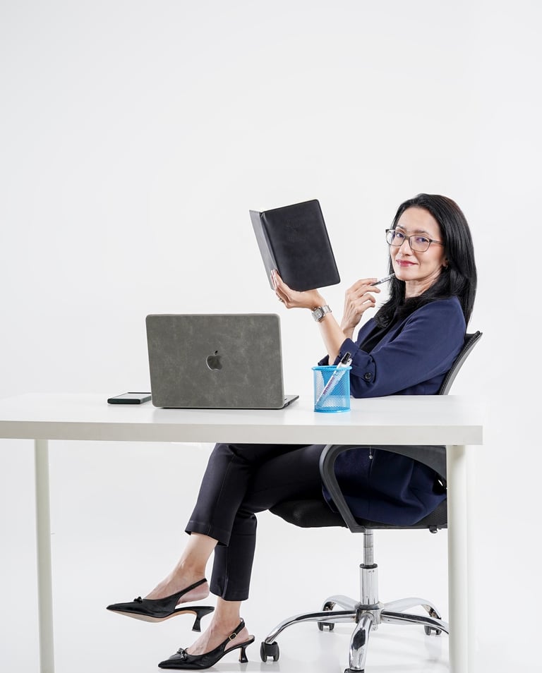 Professional businesswoman in office attire sitting at a desk with a laptop and holding a black notebook.