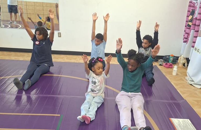 Young children sitting on a purple gym mat stretching their arms up during a group fitness class.