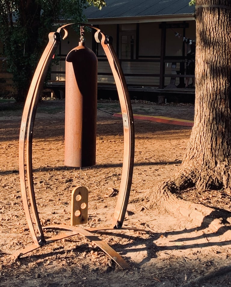 Rustic metal garden bell sculpture hanging on a curved steel frame in a sunset outdoor yard setting.