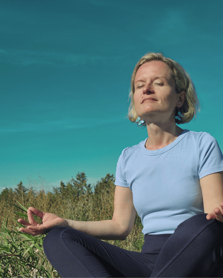 Laura in a meditation lotus position in nature, surrounded by green grass and blue skies.