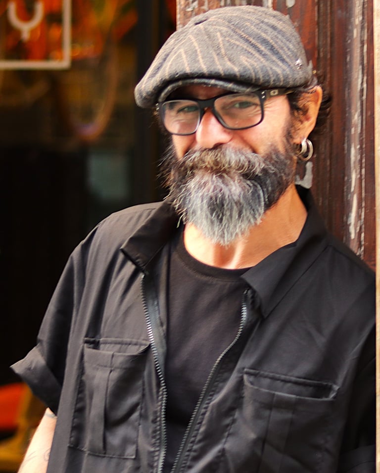 Smiling gray-bearded man in glasses and flat cap stands outdoors by orange URBAN sign, blue sky behind. Black shirt and jacke