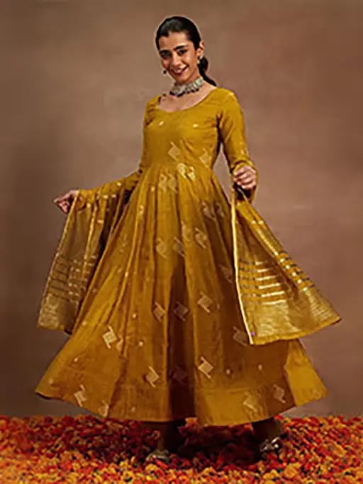 A woman posing in a mustard yellow Anarkali suit with a matching dupatta and gold embroidery.