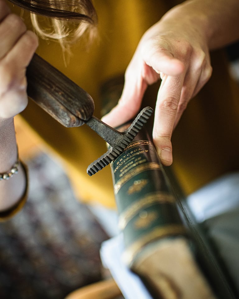Artisan using a vintage bookbinding wheel tool to emboss gold details onto a leather book spine.