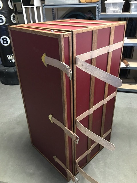 A vintage-style upright red wooden wardrobe trunk with leather straps and brass hardware in a workshop.