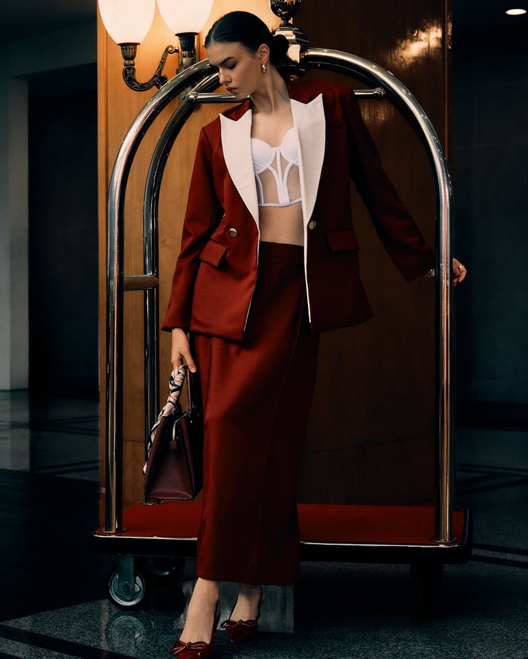 Model in a maroon blazer and maxi skirt holds a red handbag with a scarf, standing by a hotel lobby trolley.
