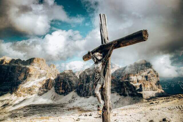 Weathered wooden crucifix with Jesus statue set against snow-capped mountains and cloudy sky.