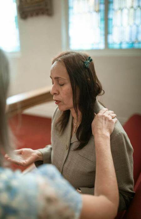 Woman with eyes closed and hands raised in prayer, with a hand placed gently on her shoulder.