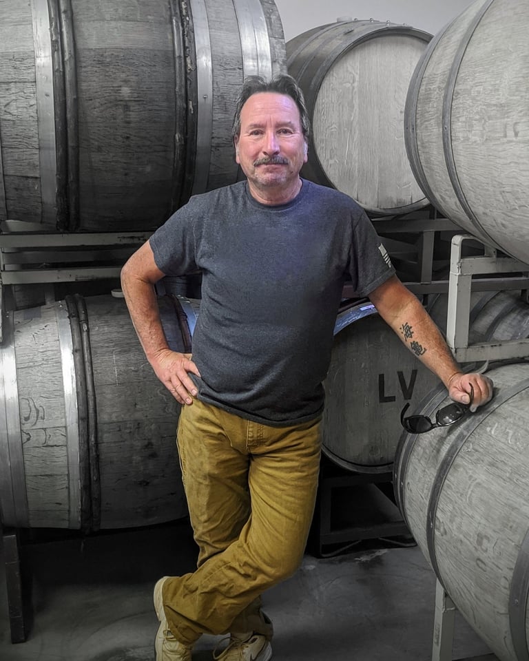 A man in a grey shirt leans against a wine barrel.