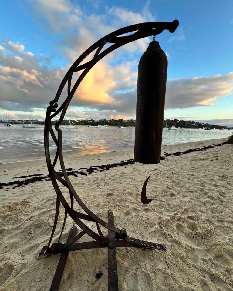 Rustic metal bell sculpture on a sandy beach at sunset with boats in the coastal bay.