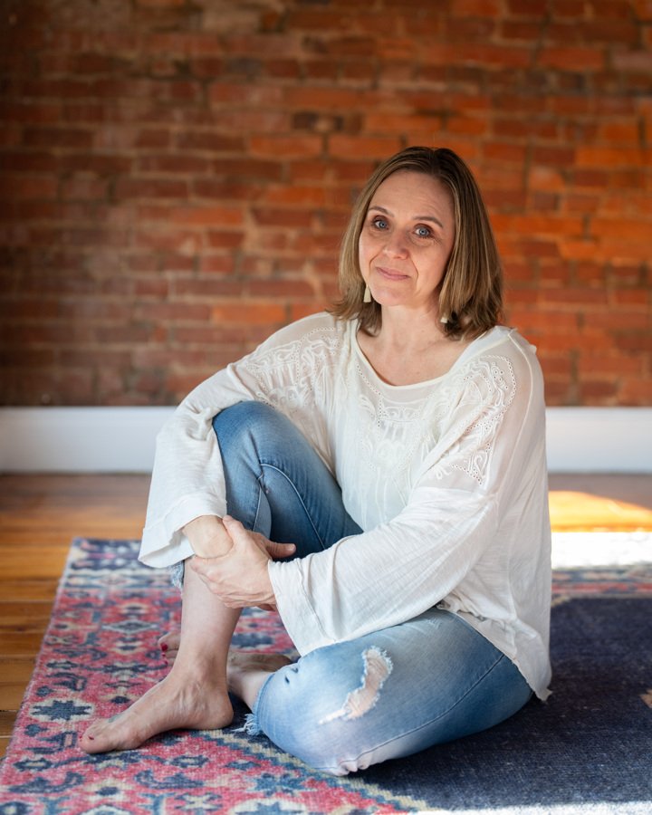 Woman sitting indoors on a rug in front of a brick wall.