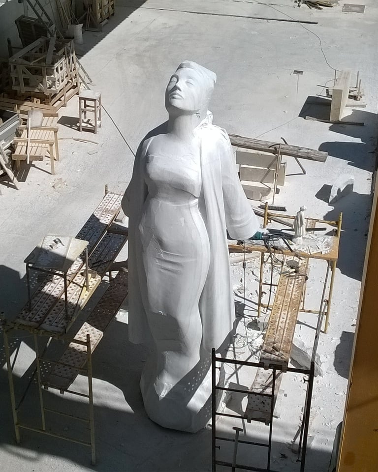 A large white marble statue of a standing woman inside a stone carving workshop with scaffolding.