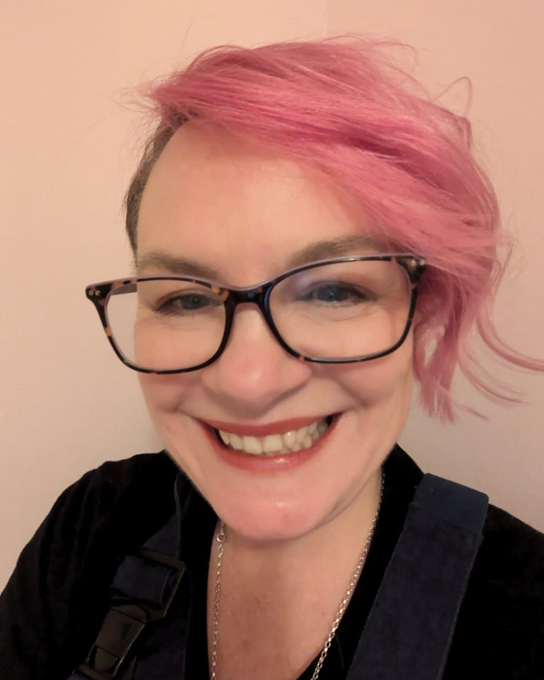 Jo de Serrano from Order with Chaos with pink hair and tortoiseshell glasses wearing blue overalls.