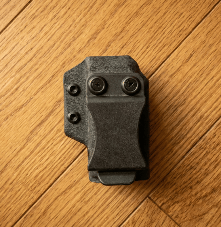 Black Kydex IWB magazine carrier with belt clip on a light oak wood background.