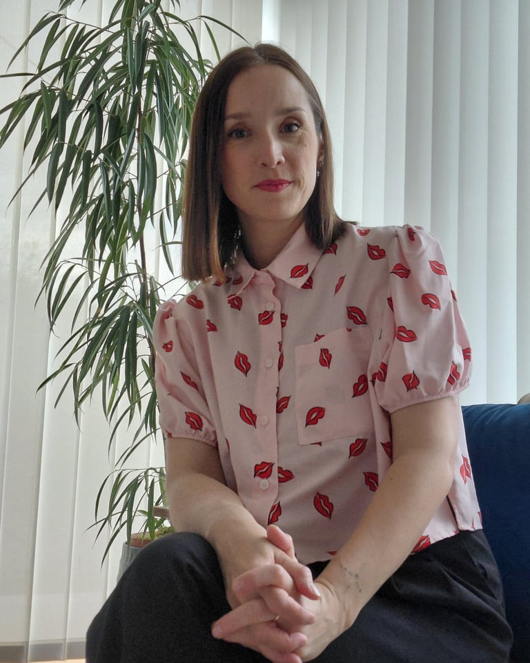 A woman wearing a pink button-up shirt with red lip prints sits next to a green indoor plant.