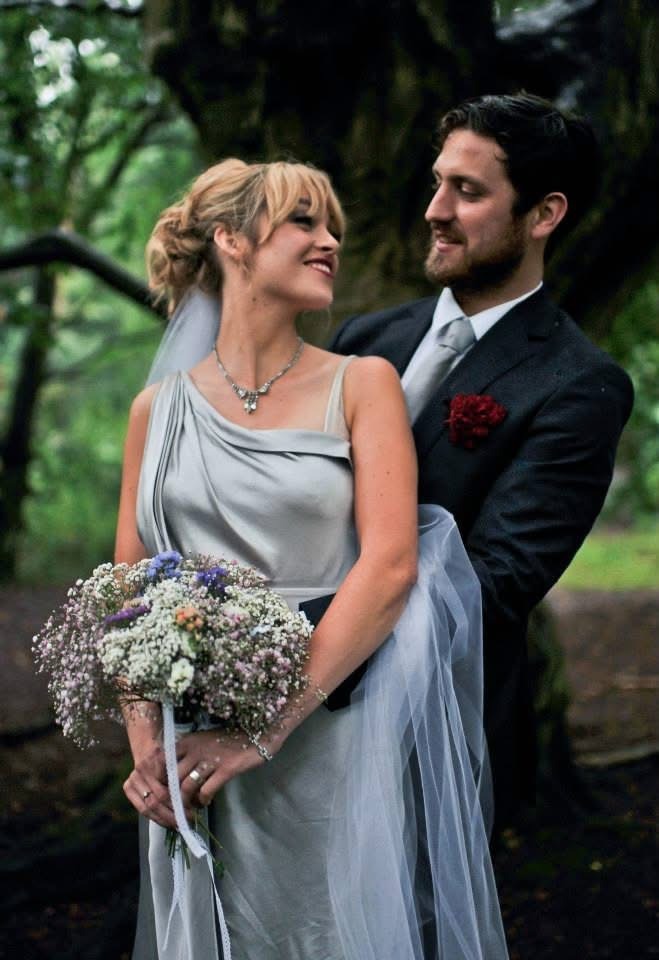 Bride in grey silk dress holding flowers in side embrace with smiling groom