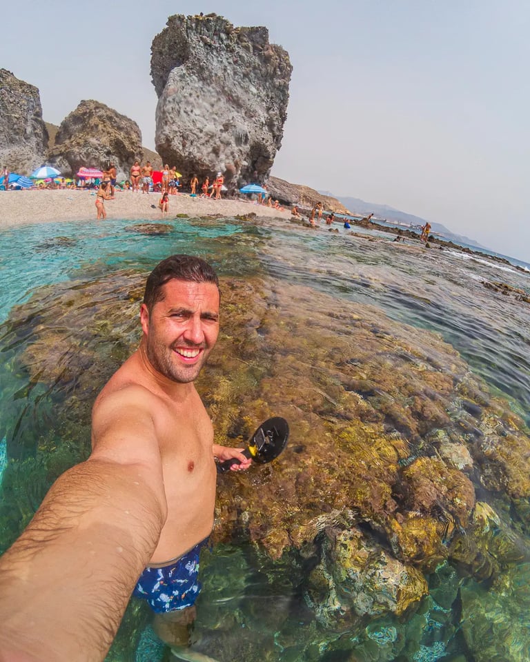 A man takes a selfie while metal detecting in clear shallow water at a crowded tropical rocky beach.