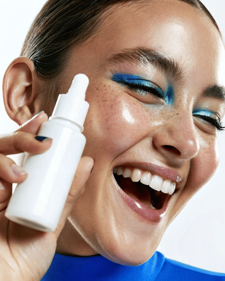Smiling woman with blue eyeshadow holding a white facial serum dropper for glowing skin care.
