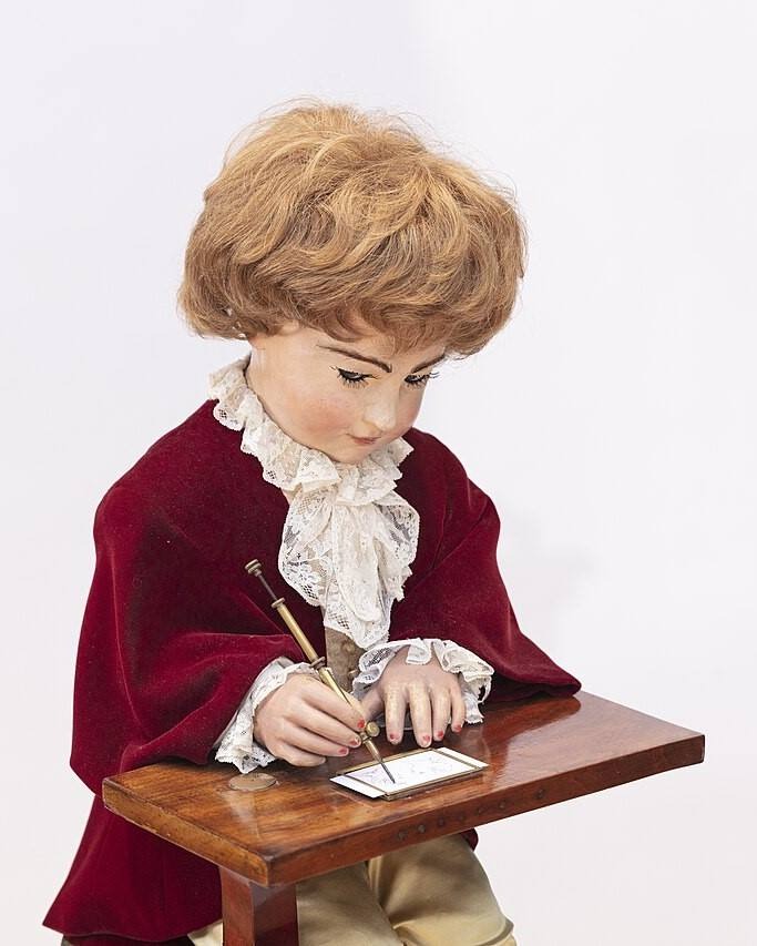 Vintage mechanical writing automaton doll in a red velvet coat sitting at a wooden desk.