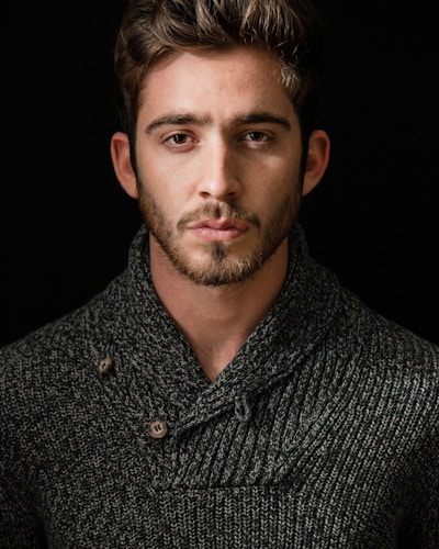A handsome man with a beard wearing a grey knit shawl collar sweater against a black background.