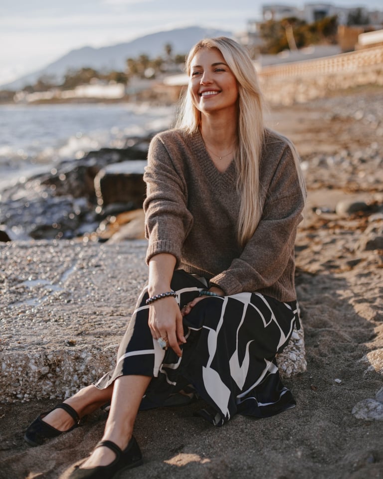Smiling blonde woman in a brown v-neck sweater and patterned skirt sitting on a beach at sunset. 