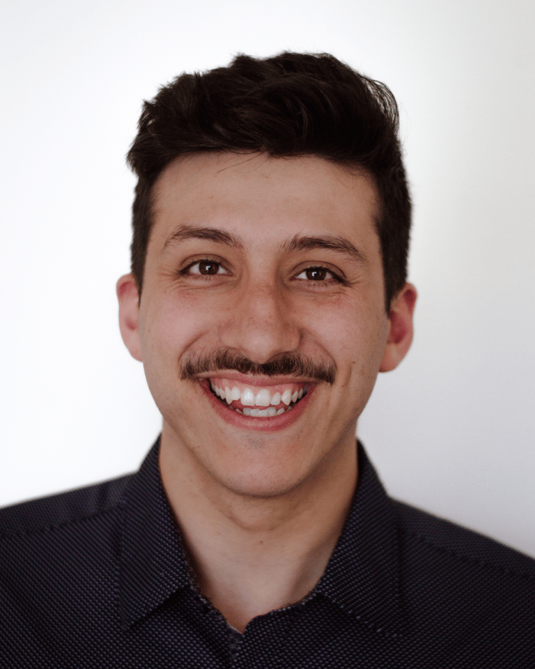 Smiling man with dark hair and mustache against a plain white background, wearing a dark shirt. 