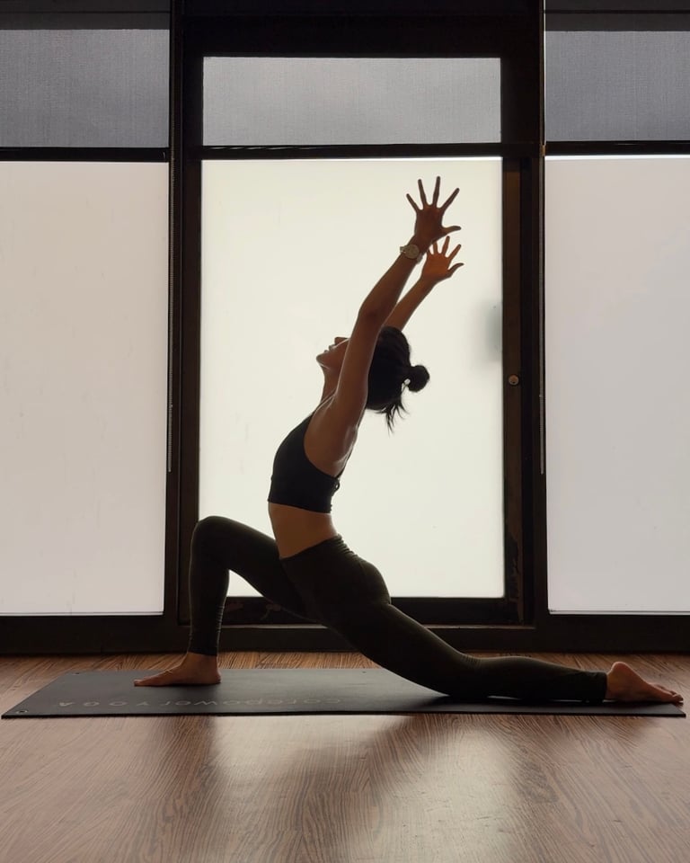 A woman practices a yoga low lunge pose on a mat in front of a window.