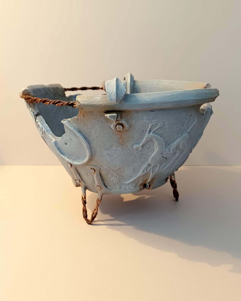 A fragmented, round bowl-like sculpture adorned with symbols and letters, finished in blue-grey hues