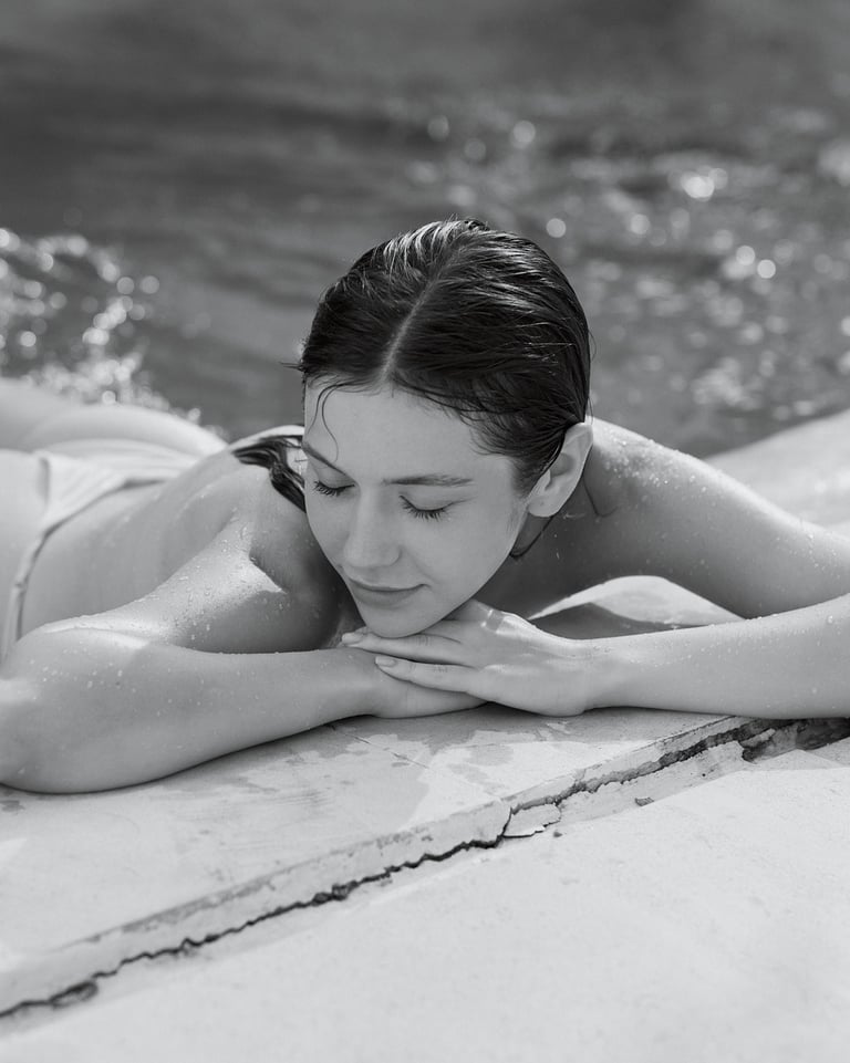 Black-and-white portrait of Anna lying by the pool edge with her upper body uncovered, resting her head on her arms.
