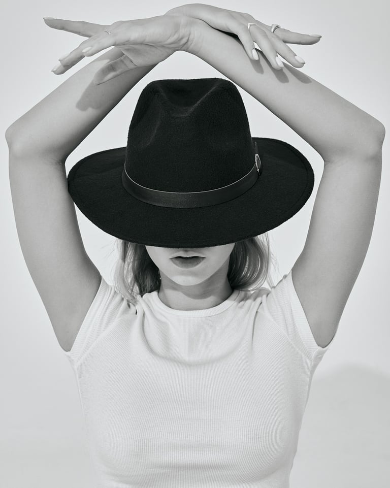 Black-and-white portrait of Kat wearing a hat and white crop top, posing with arms raised above her head.